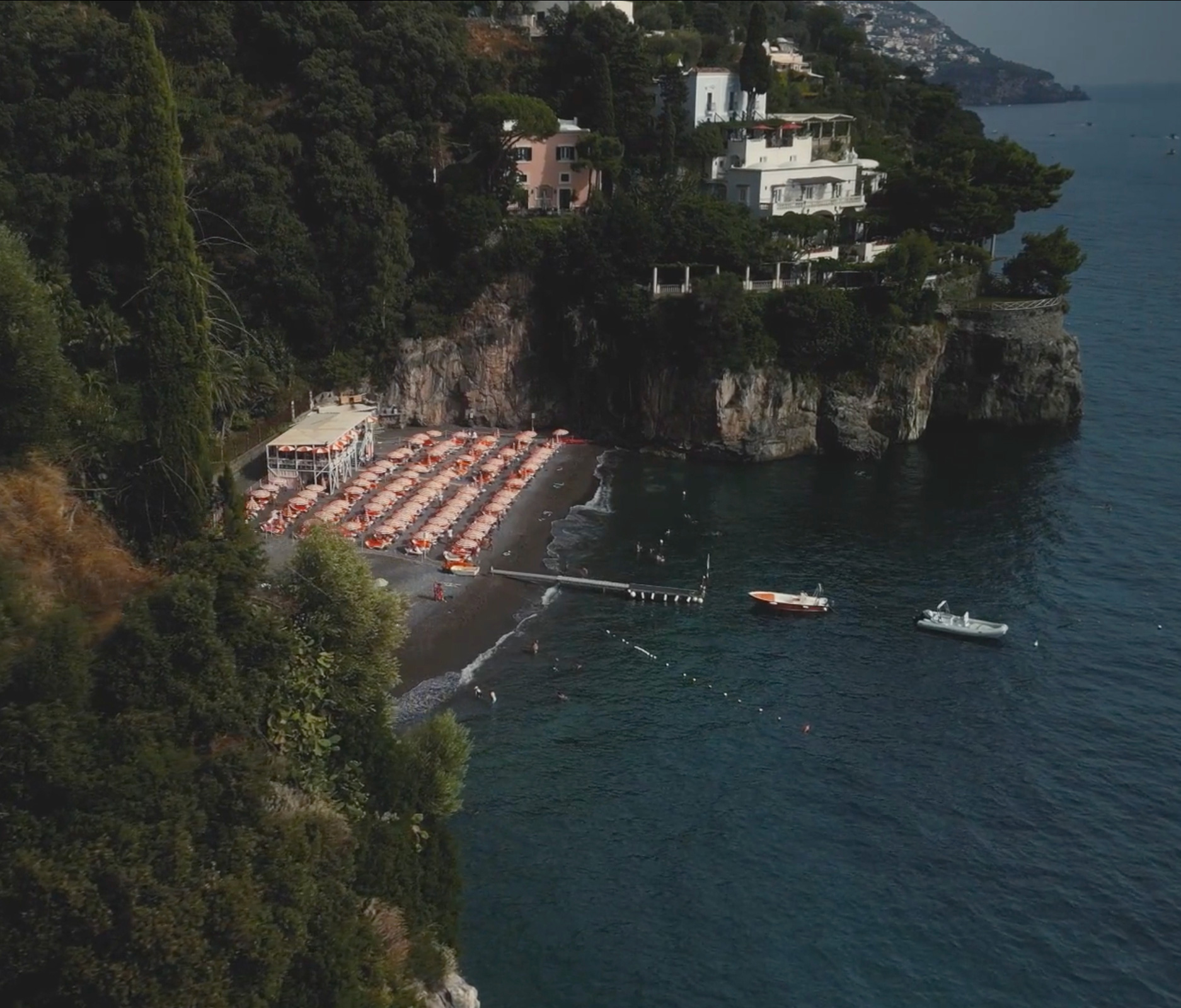 Aerial view of a secluded beach along the Amalfi Coast, featuring orange sun loungers and umbrellas, crystal-clear water, and boats anchored offshore.