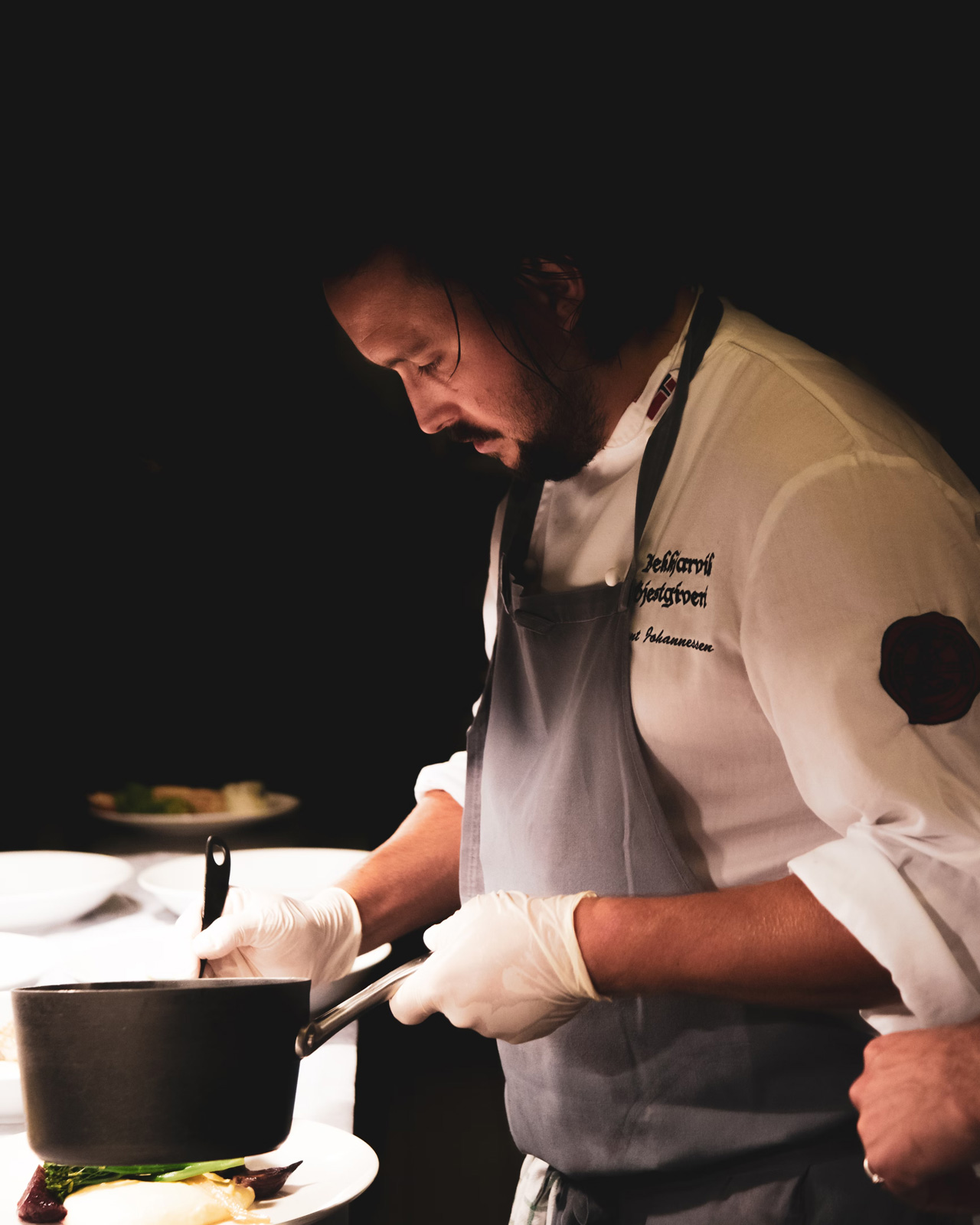 Focused chef preparing a dish in a professional kitchen, wearing white gloves and a gray apron, with a dimly lit, refined atmosphere.